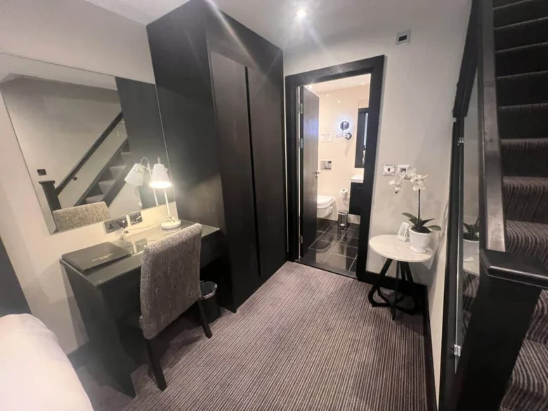 A modern hotel room with a desk and chair, a wall mirror, a wardrobe, a potted orchid on a small round table, carpeted stairs, and a view into a brightly lit bathroom.