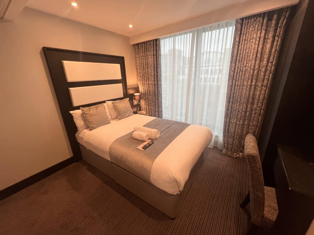 A neatly made double bed in a modern hotel room with two pillows, two towels on top, a bedside lamp, floor-to-ceiling window with sheer curtains, patterned drapes, and a desk with a chair.