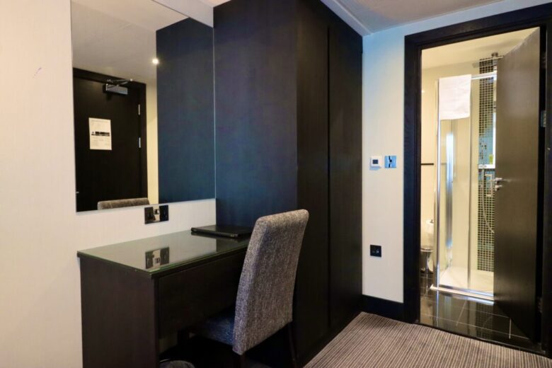 A hotel room with a desk and chair in front of a large mirror, a closed black wardrobe, and an open door revealing a bathroom with a shower and towel.