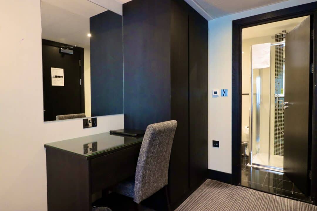 A hotel room with a desk and chair in front of a large mirror, a closed black wardrobe, and an open door revealing a bathroom with a shower and towel.