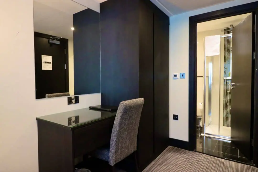 A hotel room with a desk and chair in front of a large mirror, a closed black wardrobe, and an open door revealing a bathroom with a shower and towel.