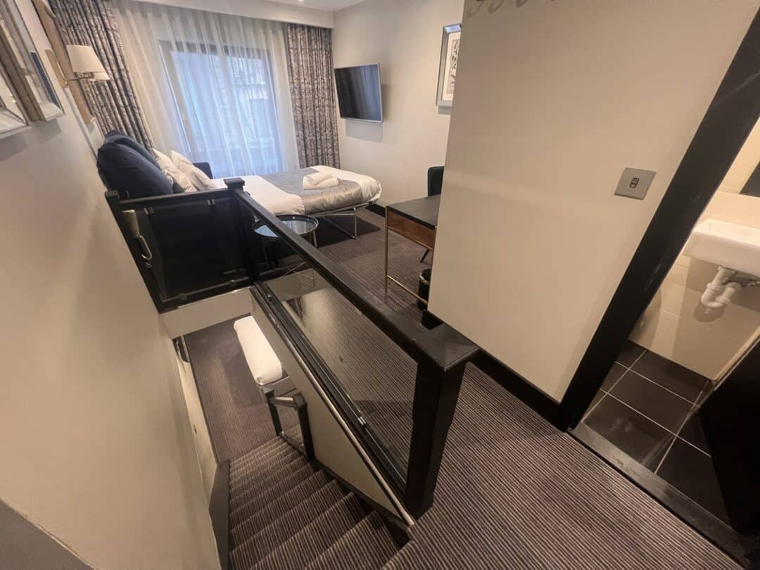 View from a staircase leading up to a modern hotel room with a bed, sofa, desk, wall-mounted TV, window with curtains, and a partial view of a bathroom sink to the right.