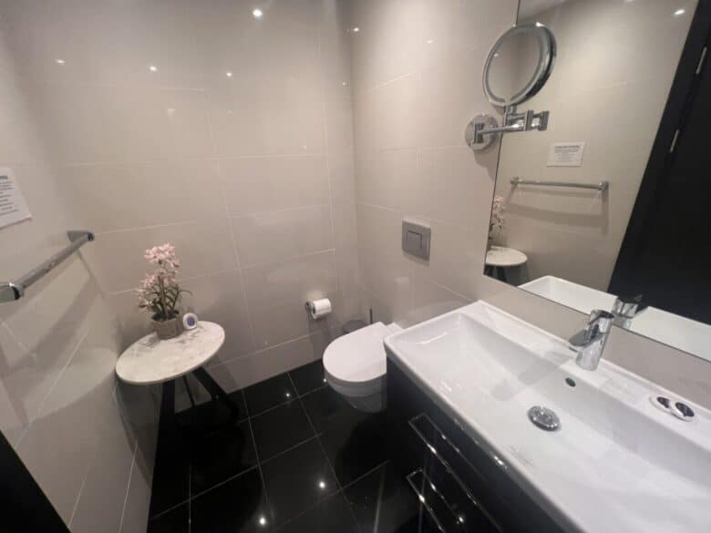 Modern bathroom with black tile floor, wall-mounted toilet, rectangular sink, round wall mirror, and a small round table with pink flowers and a rolled towel. The walls are light-colored and glossy.