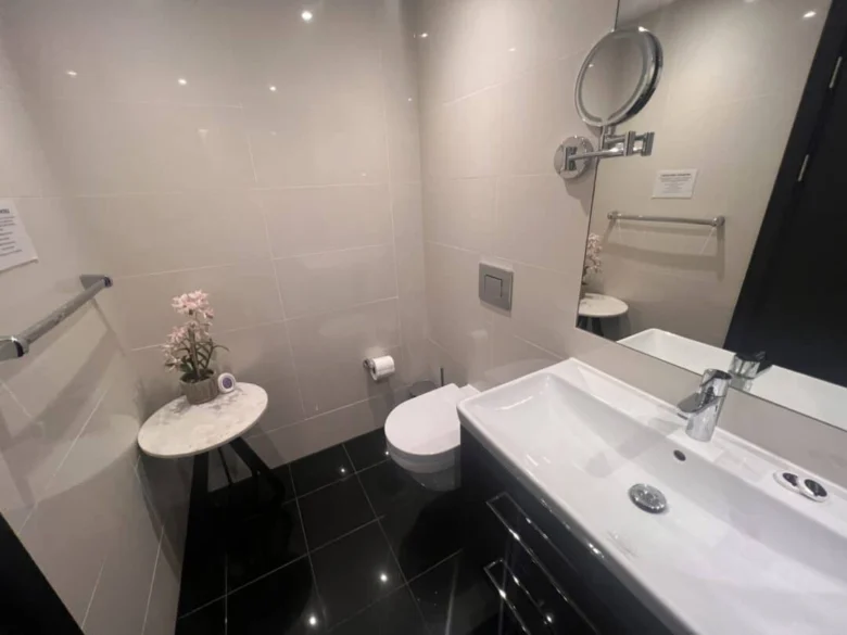 Modern bathroom with black tile floor, wall-mounted toilet, rectangular sink, round wall mirror, and a small round table with pink flowers and a rolled towel. The walls are light-colored and glossy.