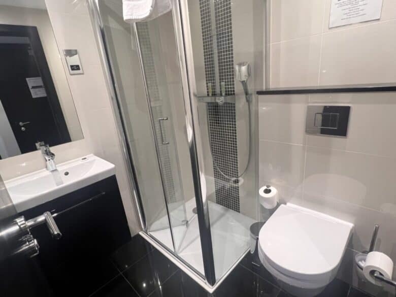 A modern bathroom with a glass-enclosed shower, wall-mounted toilet, sink with a mirror, black and white tiles, and a towel hanging above the shower. Toilet paper and toiletries are visible.