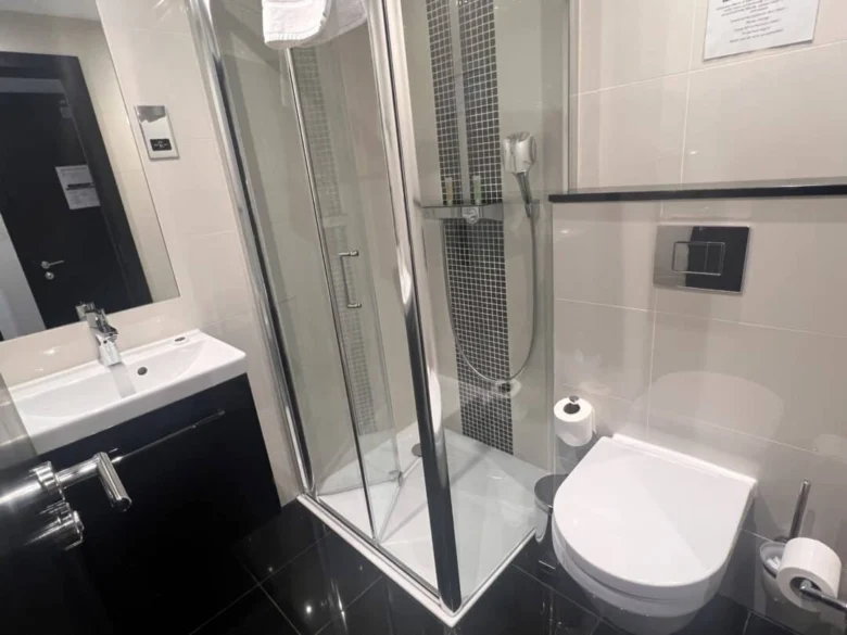 A modern bathroom with a glass-enclosed shower, wall-mounted toilet, sink with a mirror, black and white tiles, and a towel hanging above the shower. Toilet paper and toiletries are visible.