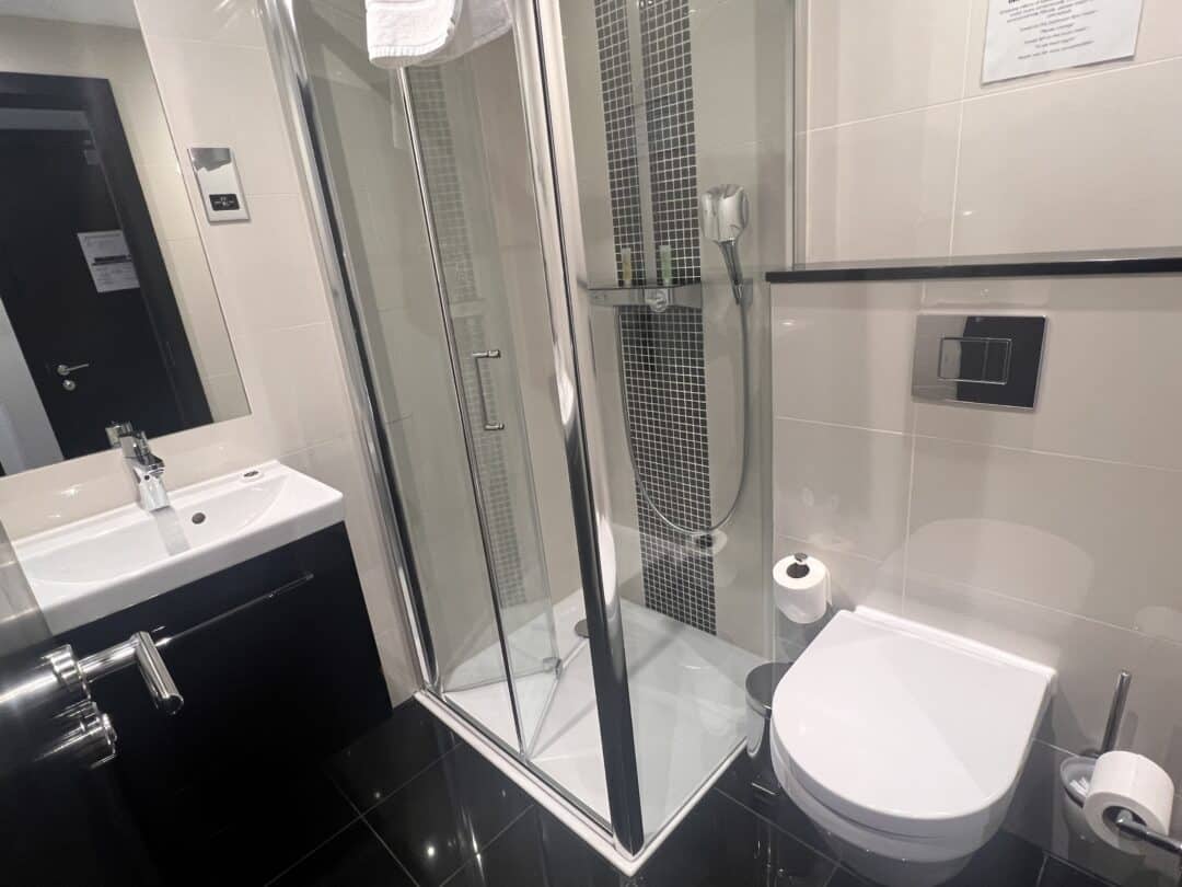 A modern bathroom with a glass-enclosed shower, wall-mounted toilet, sink with a mirror, black and white tiles, and a towel hanging above the shower. Toilet paper and toiletries are visible.