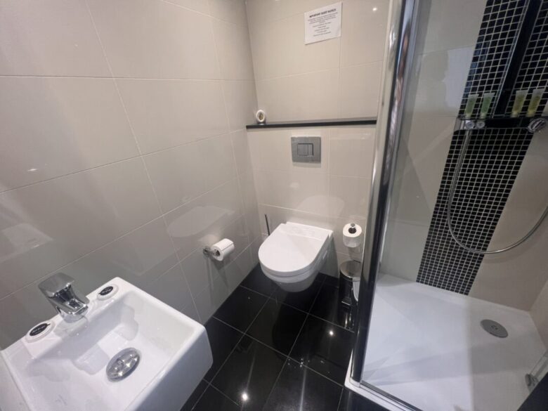 Modern bathroom with a wall-mounted toilet, a bidet, a shower with glass door and black tile accents, and a toilet paper holder; white tiled walls and glossy black tiled floor.