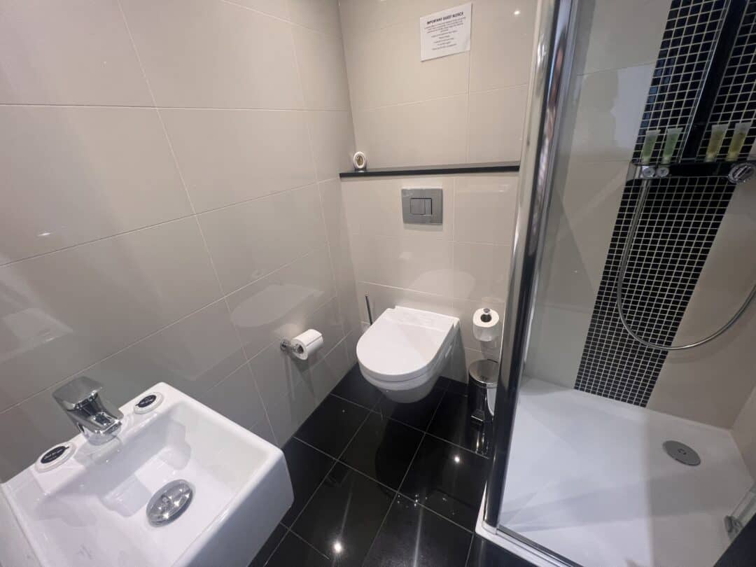 Modern bathroom with a wall-mounted toilet, a bidet, a shower with glass door and black tile accents, and a toilet paper holder; white tiled walls and glossy black tiled floor.