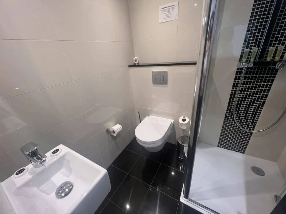 Modern bathroom with a wall-mounted toilet, a bidet, a shower with glass door and black tile accents, and a toilet paper holder; white tiled walls and glossy black tiled floor.