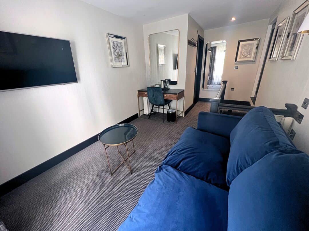 A modern hotel room with a blue sofa, round glass coffee table, wall-mounted TV, desk with a chair and mirror, and framed art on cream-colored walls. The room is carpeted and well-lit.