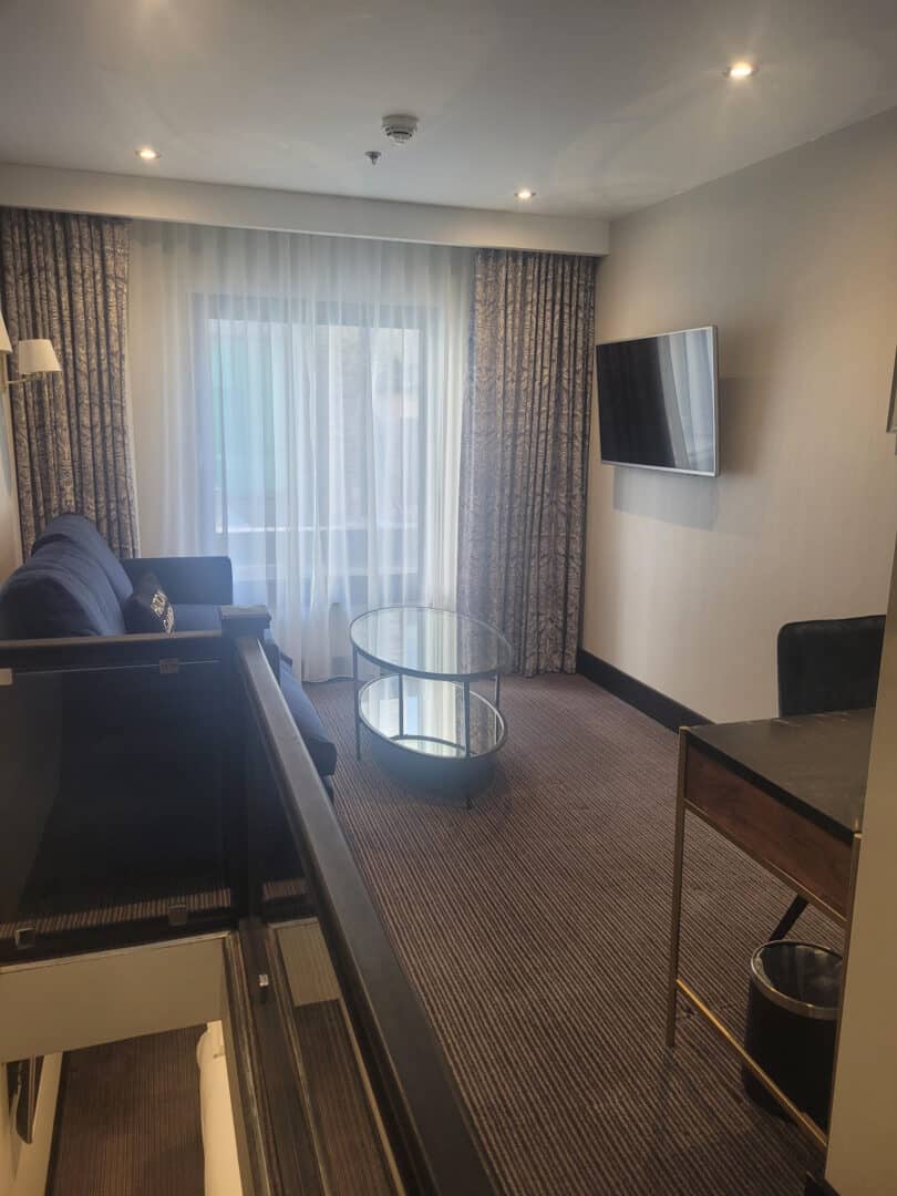 A modern hotel room with a sofa, a glass coffee table, a wall-mounted TV, a desk, and large window with curtains letting in natural light. The carpeted floor and neutral walls create a cozy atmosphere.