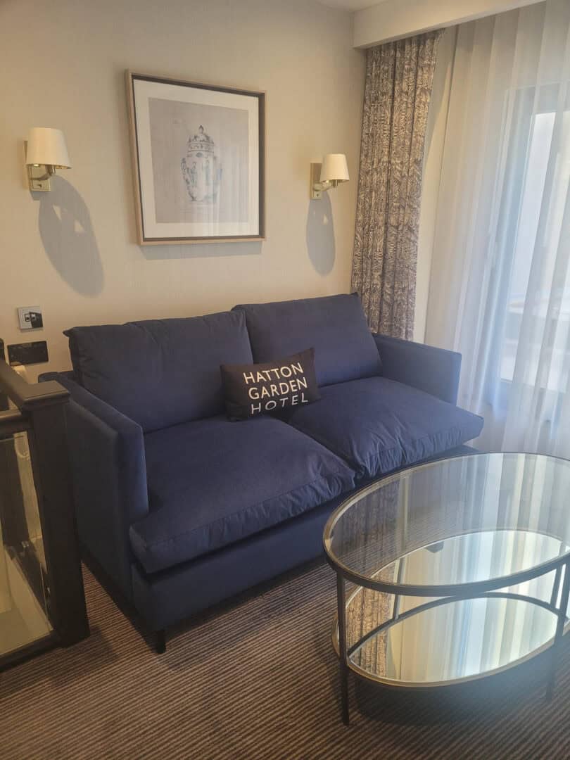 A cozy hotel room corner with a dark blue sofa, a pillow labeled "HATTON GARDEN HOTEL," a round glass coffee table, wall sconces, framed artwork, and sheer curtains letting in natural light.