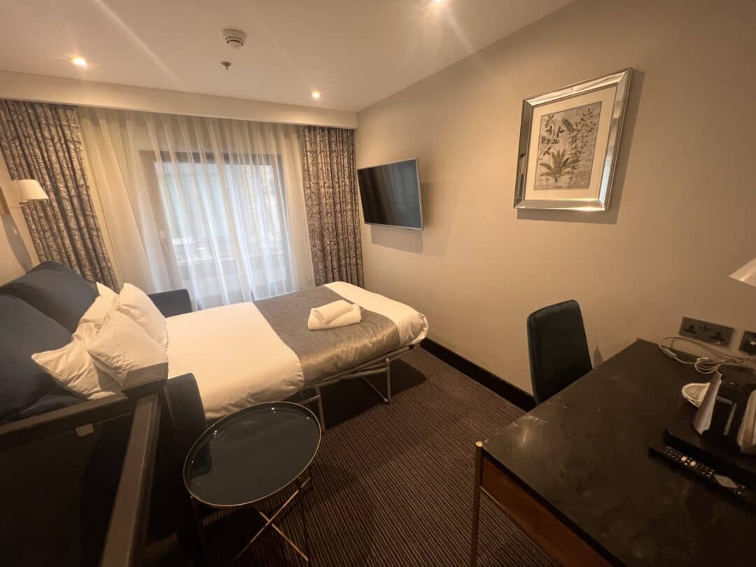 A modern hotel room with a double bed, flat-screen TV mounted on the wall, desk with a chair, framed artwork, glass side table, and large curtained window letting in natural light.