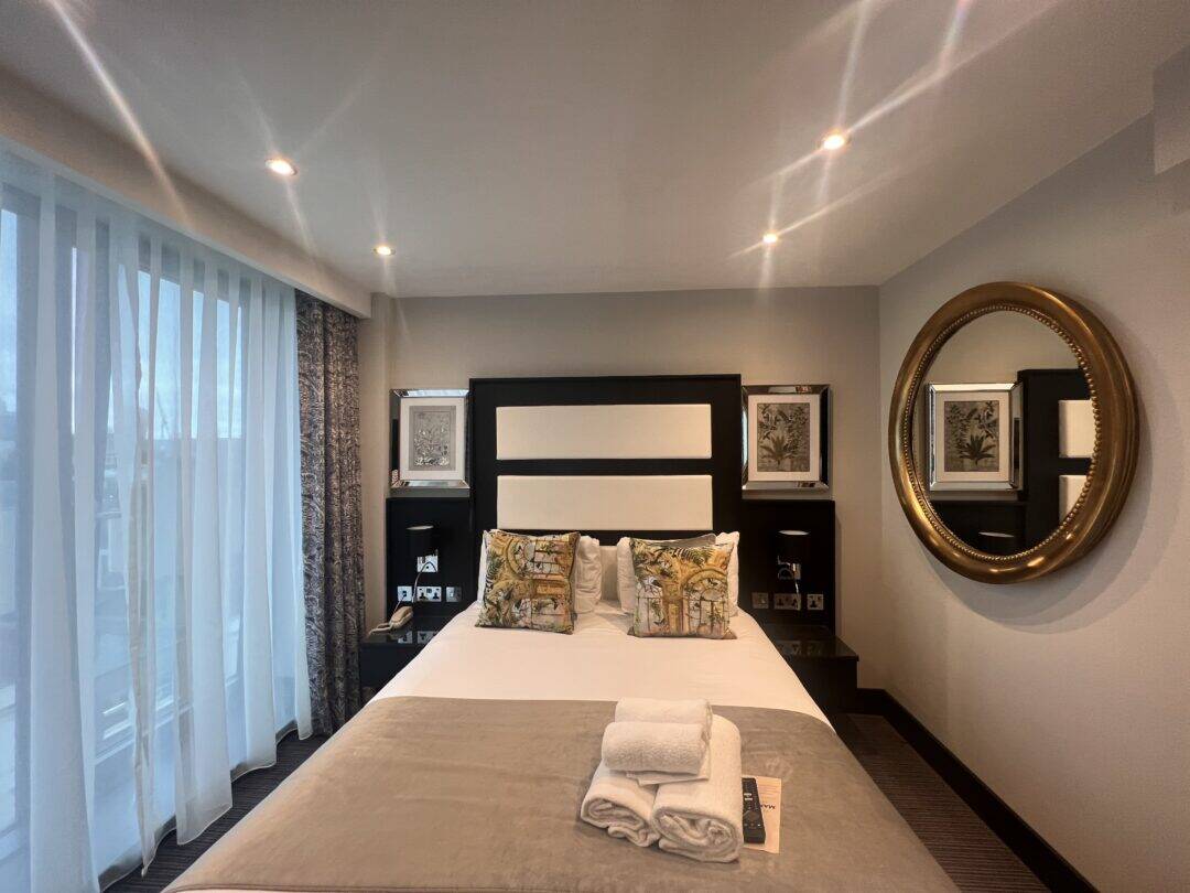 A modern hotel room with a double bed, two printed pillows, folded towels on a beige throw, large window with sheer curtains, framed art on the walls, and a round mirror reflecting part of the room.