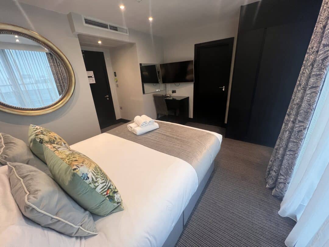 A modern hotel room with a neatly made double bed, light bedding, decorative pillows, a round mirror on the wall, a flat-screen TV, a desk, dark wardrobe, and large window with sheer curtains. Towels are folded on the bed.