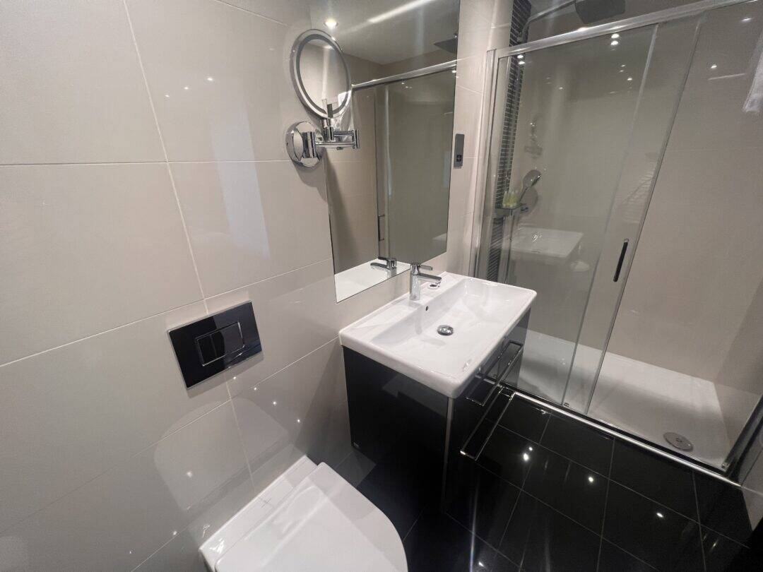Modern bathroom with a wall-mounted toilet, rectangular sink with cabinet, illuminated mirror, and a glass-enclosed shower. The room features glossy black floor tiles and light-colored wall tiles.