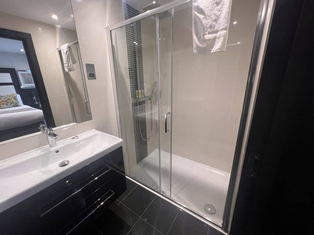 Modern bathroom with a glass-enclosed shower, a white sink with a black vanity, a towel hanging above the shower door, and a mirror reflecting a bed in the adjacent room.