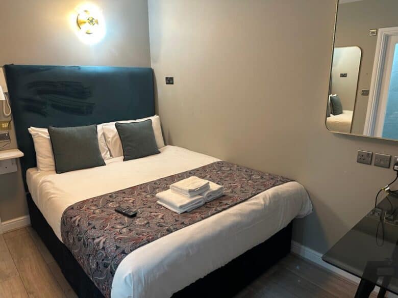 A neatly made bed with white linens, two gray pillows, and folded towels on top is in a small, modern room. A wall-mounted light, mirror, and bedside table with a remote are visible.