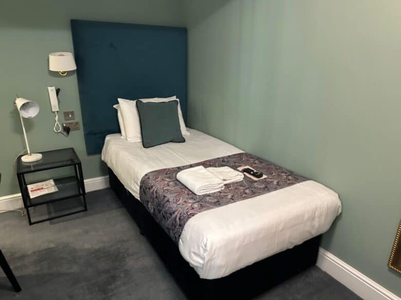 A neatly made single bed with a blue headboard, white bedding, decorative pillow, and a patterned runner. Towels and a TV remote rest on the bed. A bedside table and wall lamp are next to the bed, all in a mint green room.