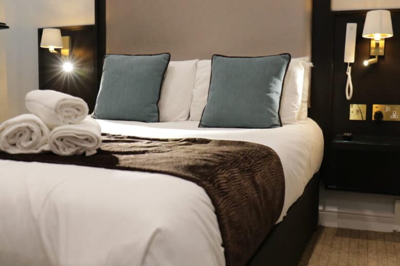 A neatly made hotel bed with white linens, two blue accent pillows, and a brown throw; three rolled white towels are placed on top. Wall lamps and a phone are mounted on the bedside wall.