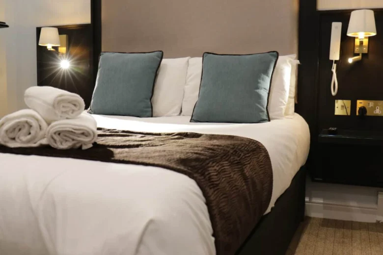 A neatly made hotel bed with white linens, two blue accent pillows, and a brown throw; three rolled white towels are placed on top. Wall lamps and a phone are mounted on the bedside wall.