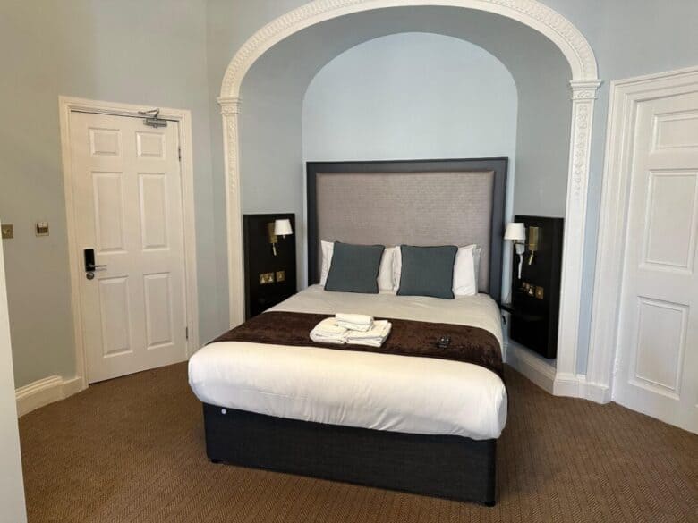 A neatly made bed with white linens, gray and teal accent pillows, and a brown throw blanket sits under an arched alcove between two doors in a light blue-walled hotel room. Towels and a TV remote are on the bed.