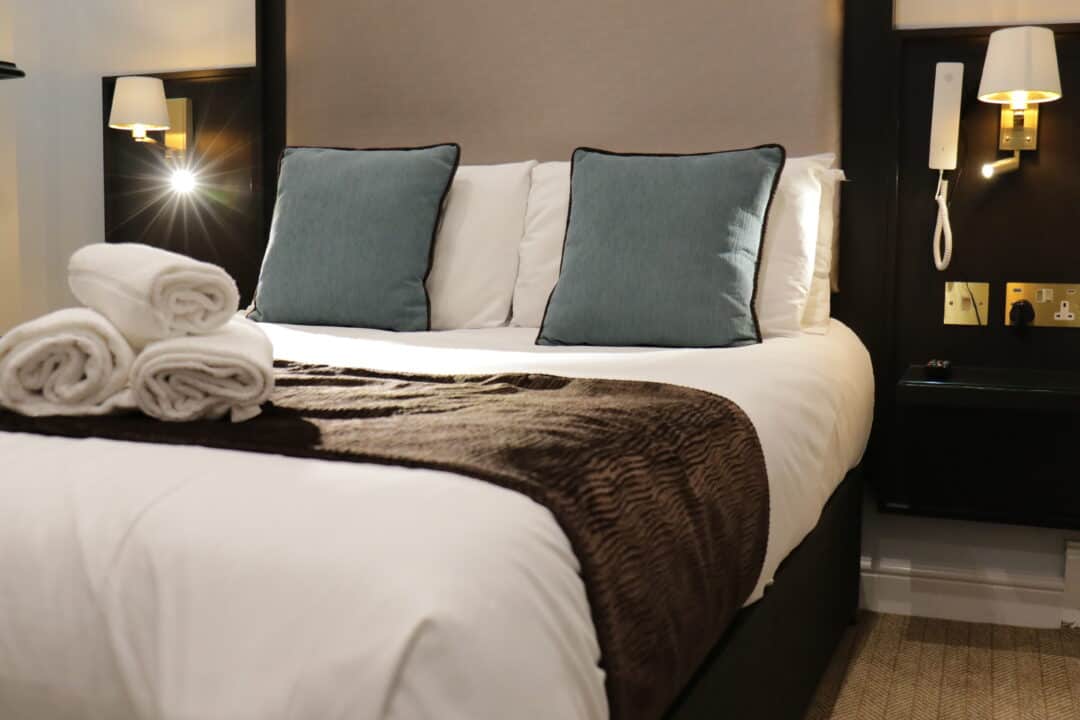 A neatly made hotel bed with white linens, two blue accent pillows, and a brown throw; three rolled white towels are placed on top. Wall lamps and a phone are mounted on the bedside wall.