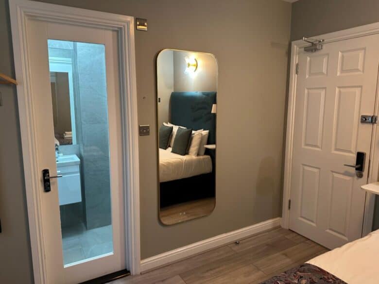 A modern bedroom with a wall mirror reflecting a bed with pillows, a white door, and an adjacent door with frosted glass revealing a bathroom sink and gray tiled walls.