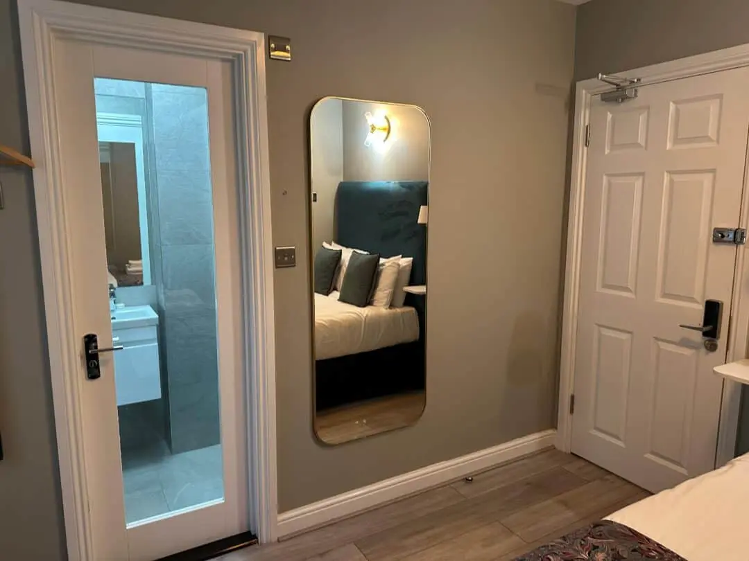 A modern bedroom with a wall mirror reflecting a bed with pillows, a white door, and an adjacent door with frosted glass revealing a bathroom sink and gray tiled walls.
