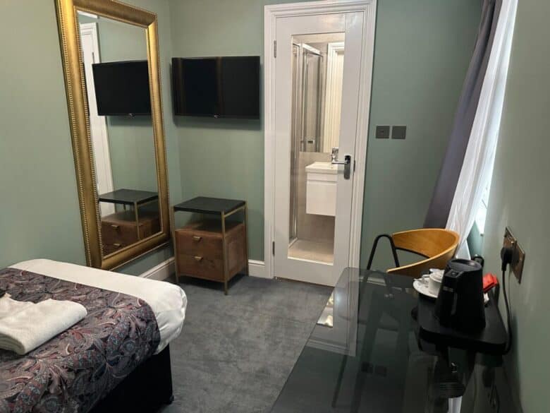 A small, tidy hotel room with a single bed, a large gold-framed mirror, two nightstands, a wall-mounted TV, a desk with a kettle and cups, a wooden chair, and an en-suite bathroom visible through an open door.