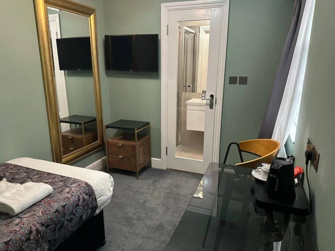 A small, tidy hotel room with a single bed, a large gold-framed mirror, two nightstands, a wall-mounted TV, a desk with a kettle and cups, a wooden chair, and an en-suite bathroom visible through an open door.
