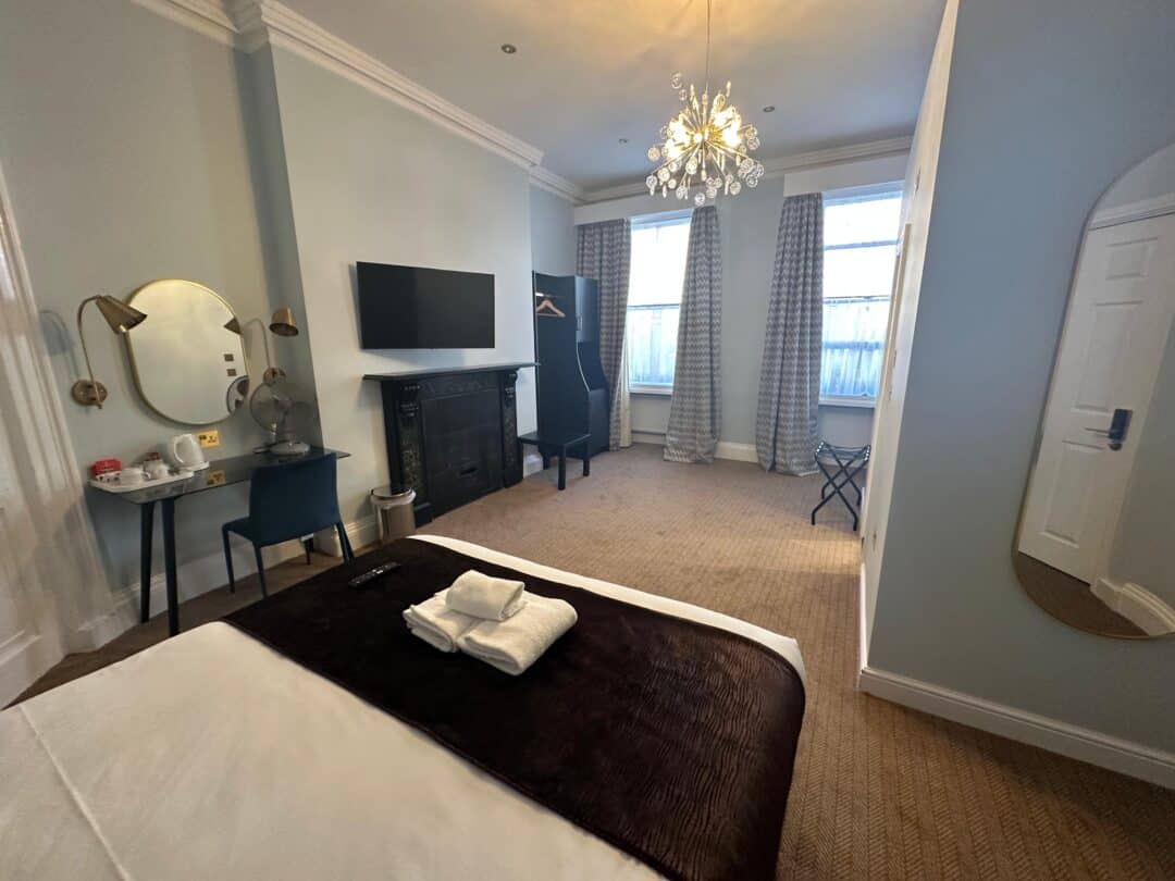 A tidy, modern bedroom with a double bed, folded towels, wall-mounted TV, desk with vanity mirror and chair, wardrobe rail, large windows with curtains, and a chandelier-style light fixture.
