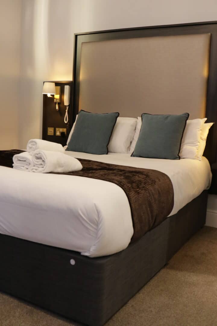 A neatly made double bed with white sheets, a brown throw blanket, and four pillows, two of which are blue. Two rolled white towels are placed on the bed. A wall lamp and telephone are mounted on the headboard.