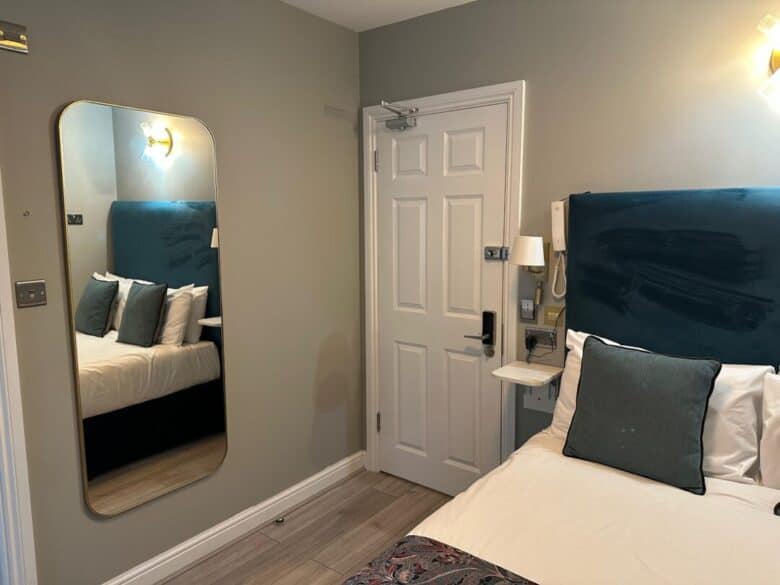A small, modern hotel room with a single bed, teal headboard, and gray pillows. A tall mirror is on the wall beside the bed, and a white door with a keypad lock is visible. Light wooden floor and muted wall colors.