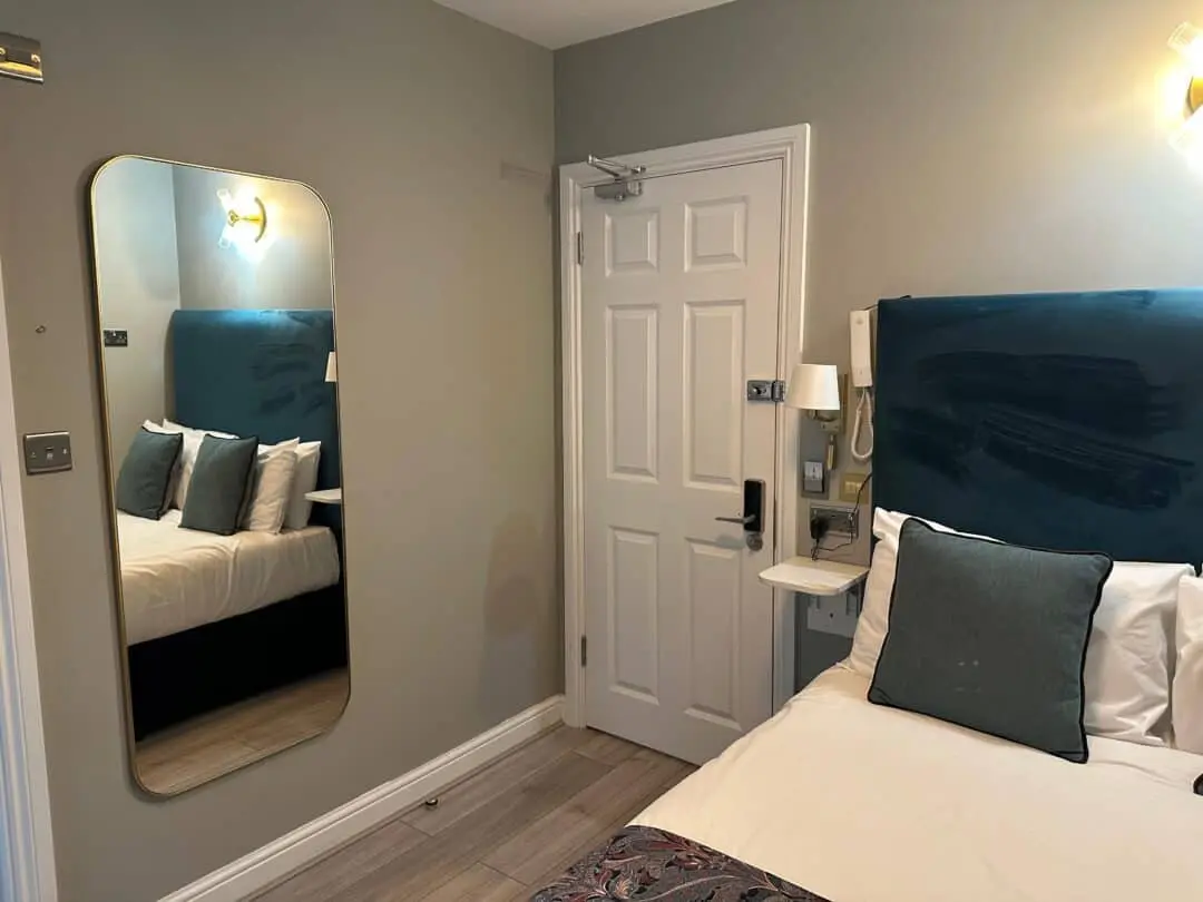 A small, modern hotel room with a single bed, teal headboard, and gray pillows. A tall mirror is on the wall beside the bed, and a white door with a keypad lock is visible. Light wooden floor and muted wall colors.