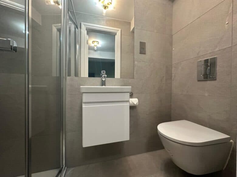 A modern bathroom with gray tiled walls, a wall-mounted sink with a mirror above it, a floating toilet, and a glass shower enclosure on the left. A light fixture is reflected in the mirror.