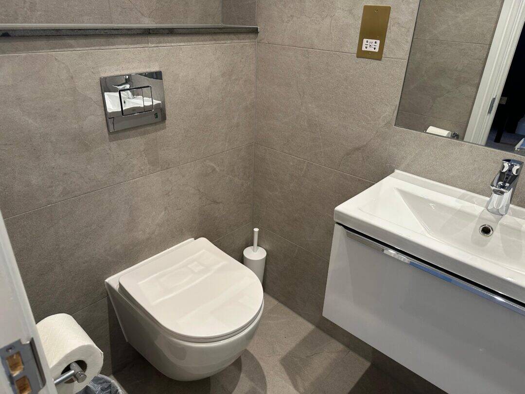 Modern bathroom with gray tiled walls, a wall-mounted toilet, a white toilet brush, toilet paper holder, a floating white sink with a chrome faucet, and a partial mirror above the sink.