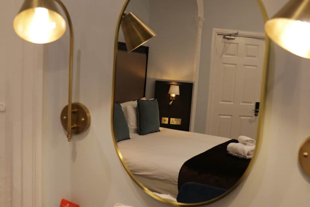 A hotel room reflected in an oval mirror, showing a neatly made bed with white linens, dark blanket, blue pillows, wall-mounted lamps, and a closed white door in the background.