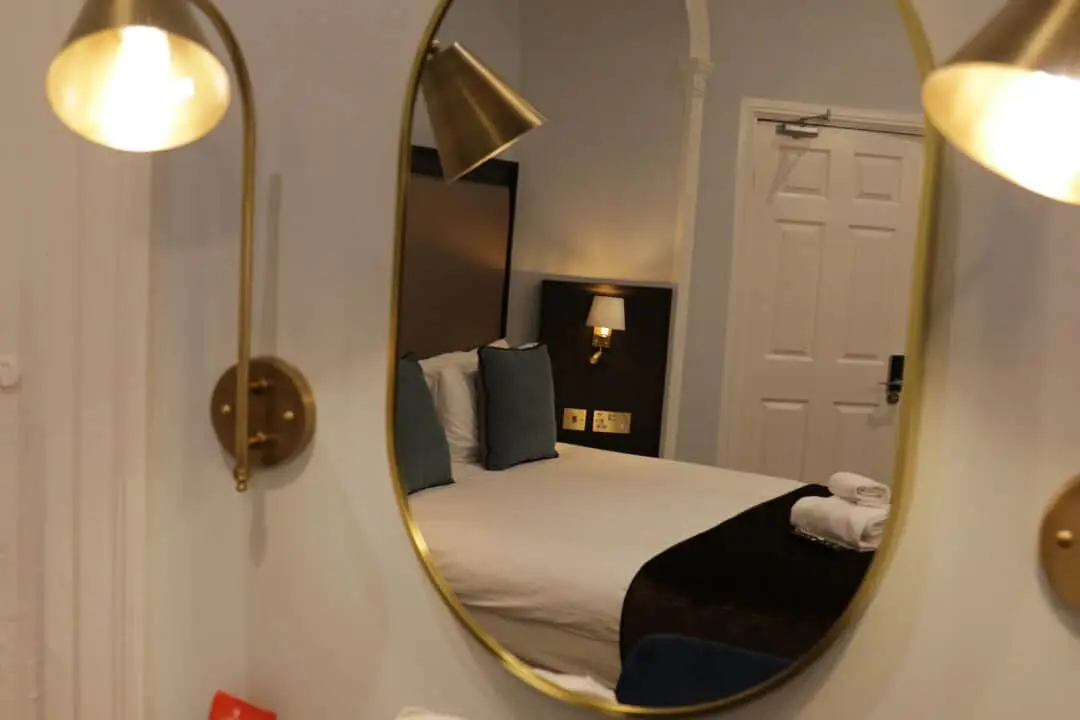 A hotel room reflected in an oval mirror, showing a neatly made bed with white linens, dark blanket, blue pillows, wall-mounted lamps, and a closed white door in the background.