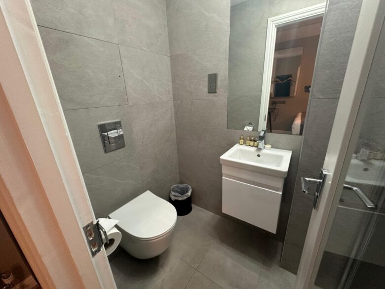 A modern bathroom with gray tiled walls and floor, featuring a wall-mounted toilet, small trash bin, floating white sink with toiletries, and a mirror above the sink. A door is partially open.