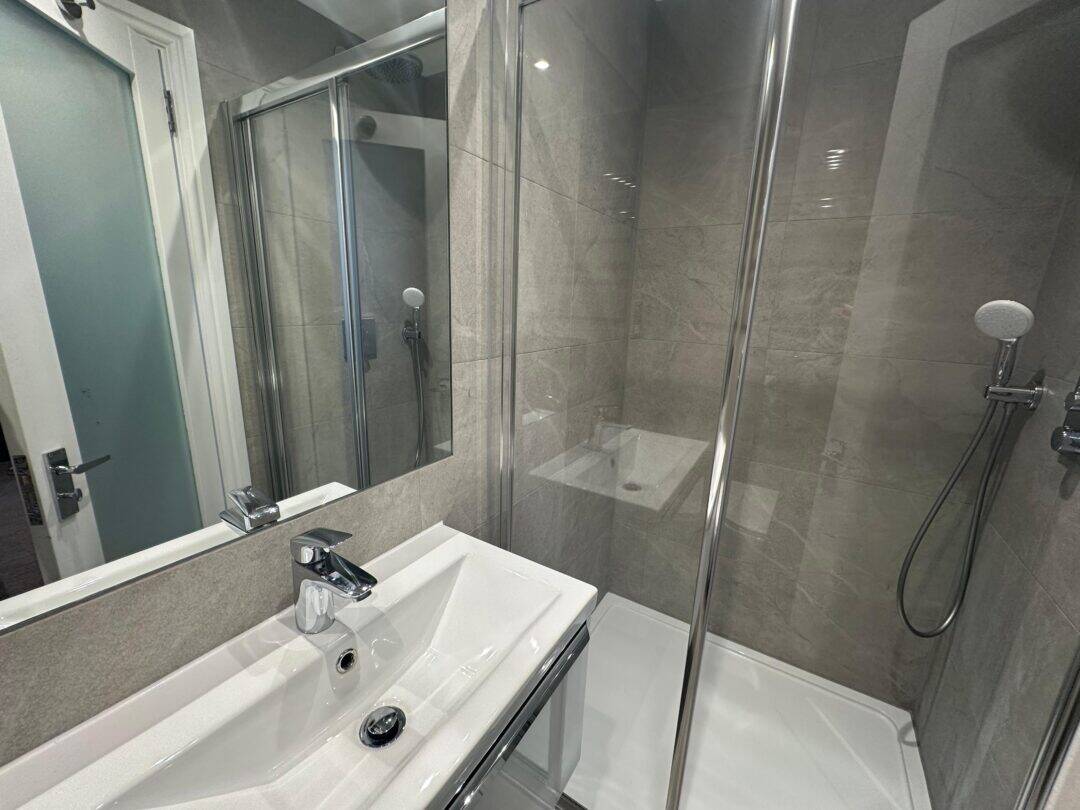 Modern bathroom with a white sink, silver faucet, glass shower enclosure, and a wall-mounted showerhead. Grey tiled walls are reflected in a large mirror above the sink.