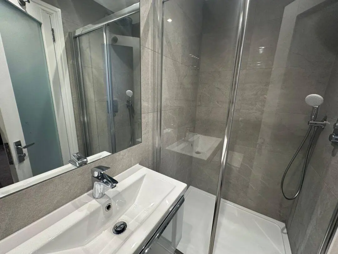 Modern bathroom with a white sink, silver faucet, glass shower enclosure, and a wall-mounted showerhead. Grey tiled walls are reflected in a large mirror above the sink.