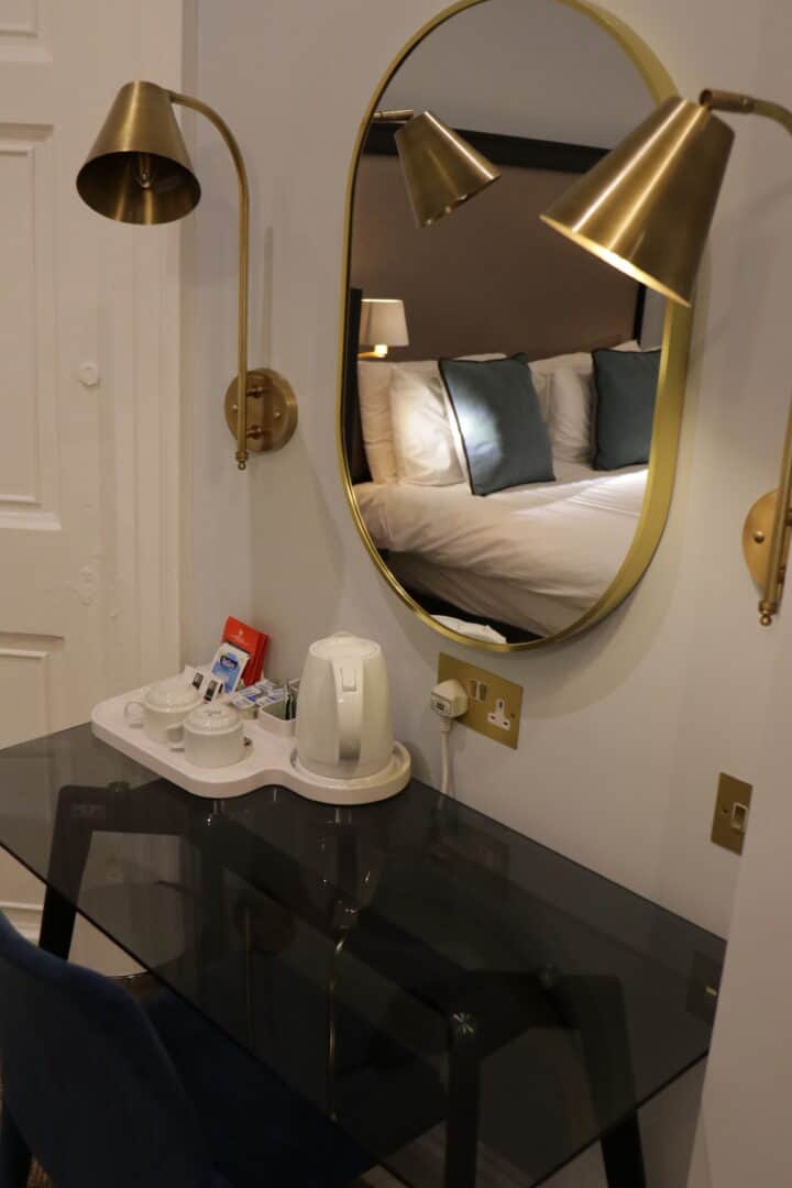 A glass desk with a tea set and electric kettle sits below a round mirror, which reflects a neatly made bed with white linens and blue pillows. Brass wall lamps are mounted on either side of the mirror.