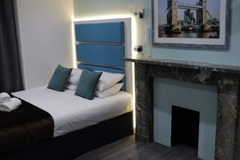 A modern hotel room with a double bed featuring blue headboard and pillows, a marble fireplace, a bedside lamp, and a framed picture of Tower Bridge on the wall above the fireplace.