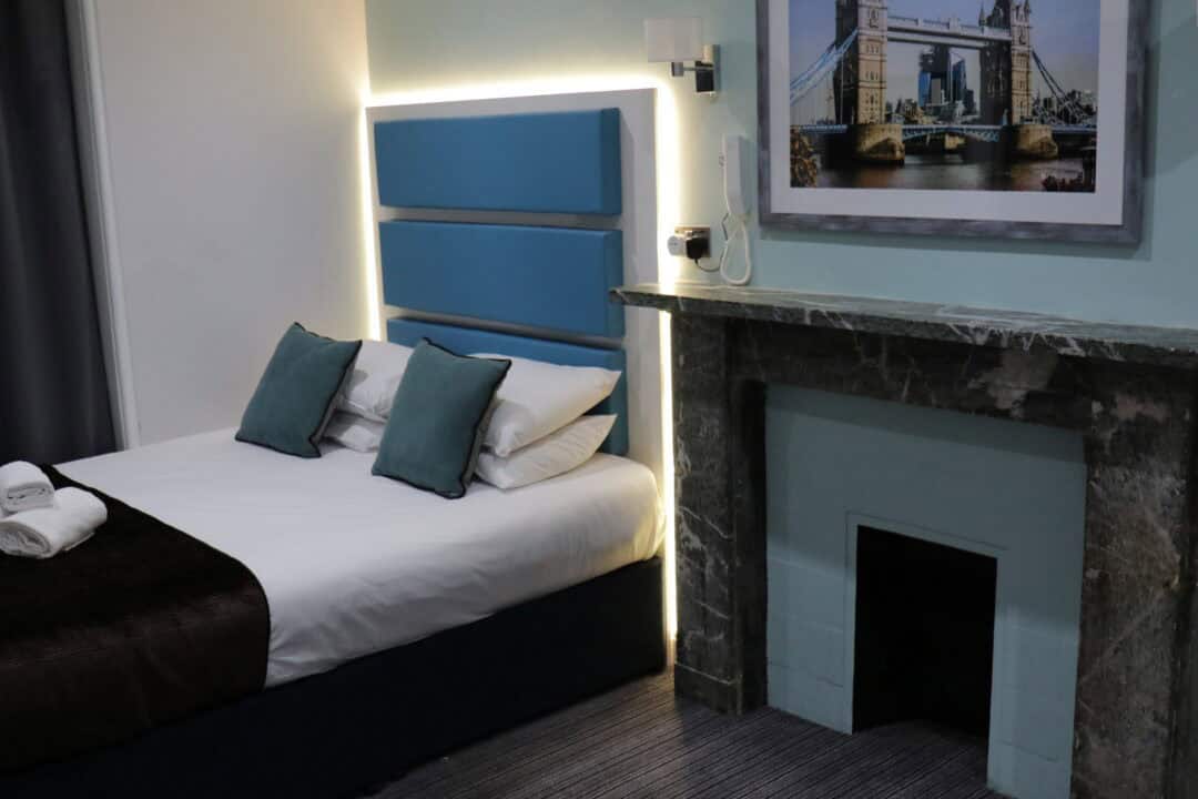 A modern hotel room with a double bed featuring blue headboard and pillows, a marble fireplace, a bedside lamp, and a framed picture of Tower Bridge on the wall above the fireplace.