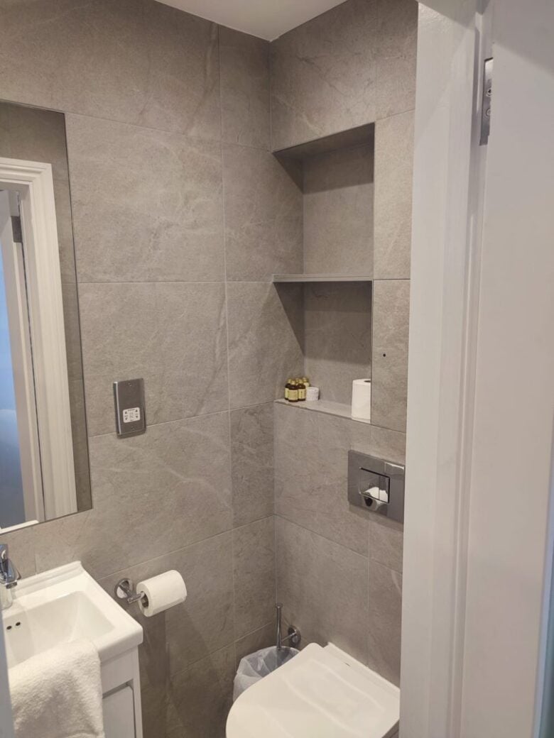 A modern bathroom with gray tiled walls, a white sink, a wall-mounted toilet, built-in shelves holding toiletries and toilet paper, a toilet roll holder, and a small trash bin.