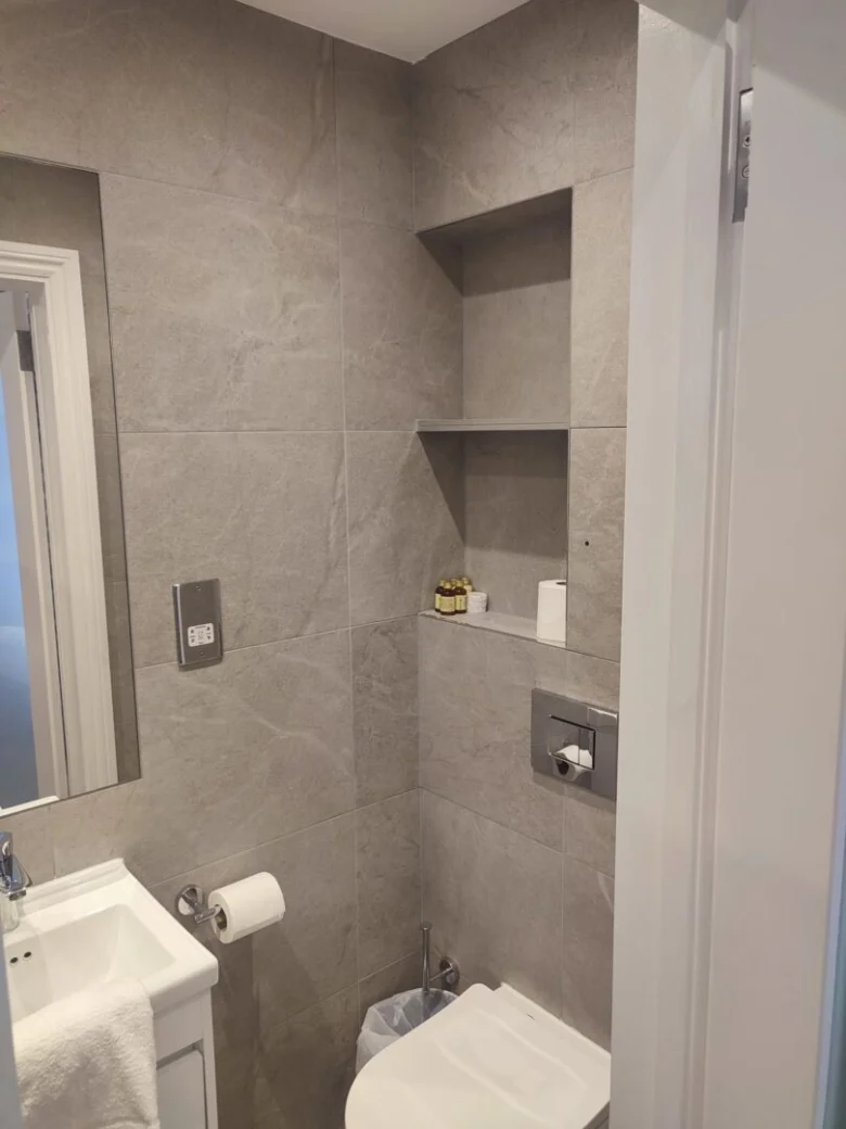 A modern bathroom with gray tiled walls, a white sink, a wall-mounted toilet, built-in shelves holding toiletries and toilet paper, a toilet roll holder, and a small trash bin.