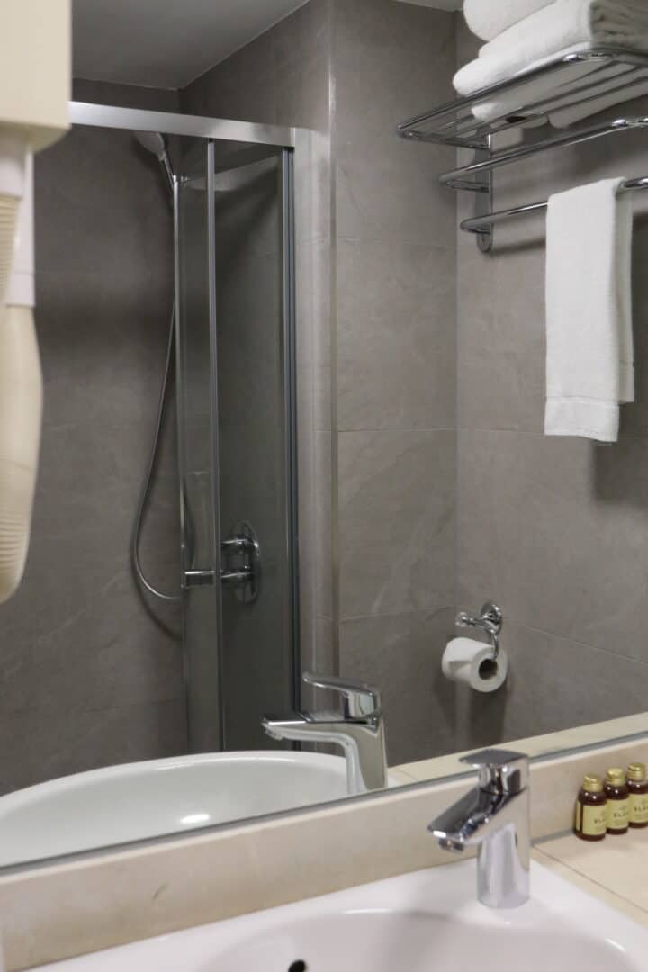 A modern bathroom with a glass shower, gray tiled walls, a white sink with a chrome faucet, a mirror, white towels on a shelf, a wall-mounted hairdryer, and toiletries on the counter.