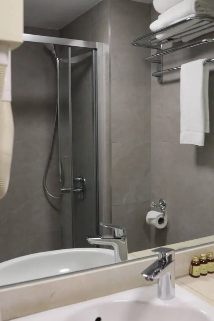 A modern bathroom with a glass shower, gray tiled walls, a white sink with a chrome faucet, a mirror, white towels on a shelf, a wall-mounted hairdryer, and toiletries on the counter.